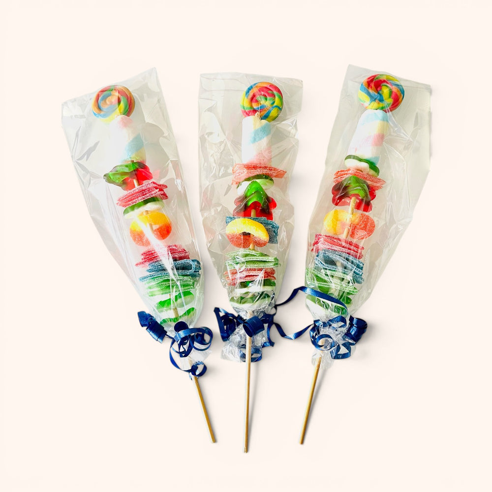 CandyCakeExplosion candy kabobs featuring colorful gummies, marshmallows, and lollipops on skewers in clear wrappers.