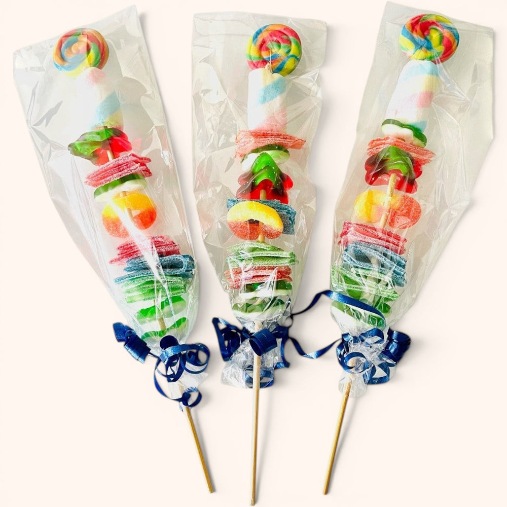 CandyCakeExplosion candy kabobs featuring colorful gummies, marshmallows, and lollipops on skewers in clear wrappers.