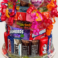 CandyCakeExplosion yellow-themed candy cake featuring assorted chocolates, Airheads, and Sour Patch Kids with ribbons.